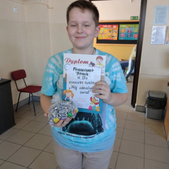 Uśmiechnięty uczeń z klasy IV z dyplomem i słodkim upominkiem, w tle sala lekcyjna