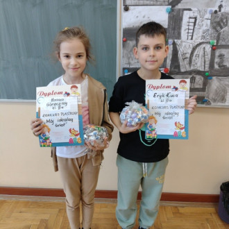 Uczennica i uczeń z klasy III z dyplomami i słodkimi upominkami, w tle sala lekcyjna