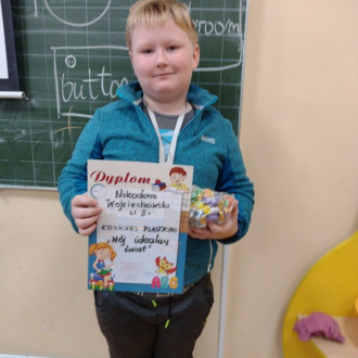 Uśmiechnięty uczeń z klasy II z dyplomem i słodkim upominkiem, w tle sala lekcyjna